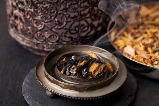 Plantation agarwood chips smouldering in a silver-rimmed amber glass mabkhara — the visual signature of a verified bakhoor chip base